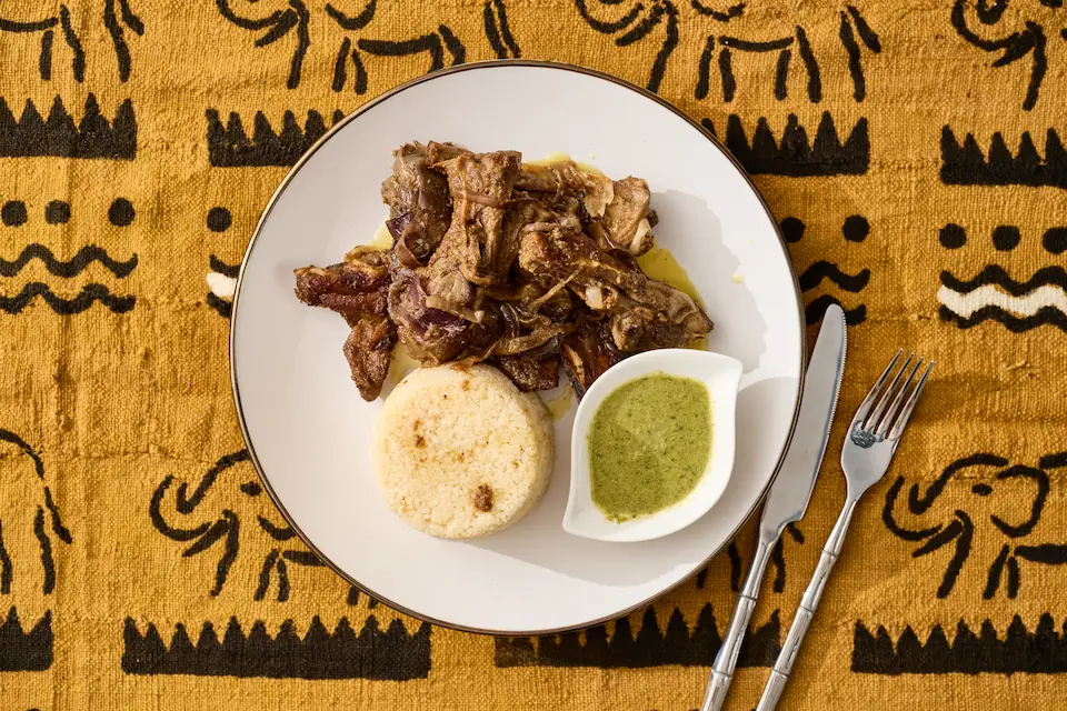 Choukouya d'agneau — morceaux d'agneau braisés aux épices ivoiriennes, servis avec attiéké frais (semoule de manioc fermentée) et sauce verte relevée, spécialité du Maquis Chic — Mauya Lyon