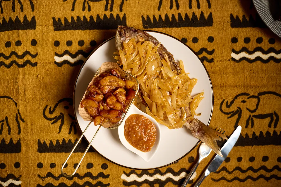 Alloco poisson — tilapia entier braisé au charbon de bois, dressage bistronomique français, servi avec alloco (bananes plantains frites ivoiriennes) et sauce tomate maison aux herbes — Mauya Lyon